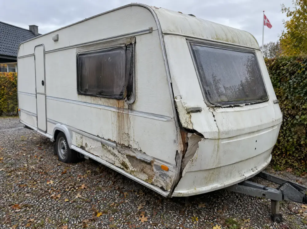 Defekt campingvogn med fugtskade og ødelagt hjørne på en gruset indkørsel